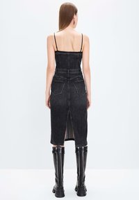 Vestido midi de mezclilla negra con tirantes finos, abertura en la parte posterior y dos bolsillos traseros, combinado con botas negras de caña alta. Textura suave.