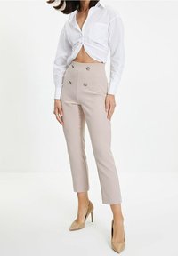 Beige op maat gemaakte broek met dubbele knoopdetails en een cropped lengte, gecombineerd met een wit overhemd met knopen. Gladde stof en getailleerd ontwerp.