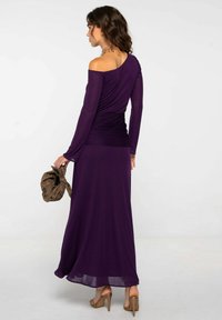 Mujer con un vestido largo morado con los hombros al descubierto, sosteniendo un bolso de mano marrón, vistiendo sandalias de tacón alto beige, de pie contra un fondo blanco.