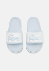 Zapatos de playa de color azul claro con suelas texturizadas, con un gran logotipo de cocodrilo embosado en una amplia correa blanca. Material suave y liso.