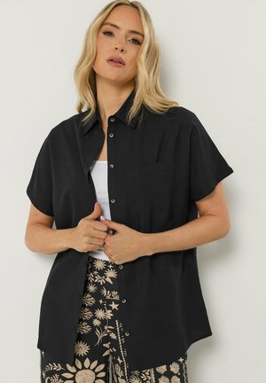 Frau mit blondem Haar, die ein locker sitzendes schwarzes kurzärmeliges Hemd mit Knöpfen über einem weißen Top und schwarzen Hosen mit Blumenmuster trägt.