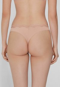 Nude-farbener Tanga mit einem gewellten Spitzenbund, der durch eine glatte Textur und ein körperbetontes Design besticht und einen minimalistischen Stil betont.