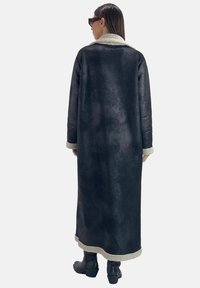 Abrigo negro de borrego con silueta larga, superficie texturizada, cuello y bajo de piel crema, combinado con botines negros hasta el tobillo.