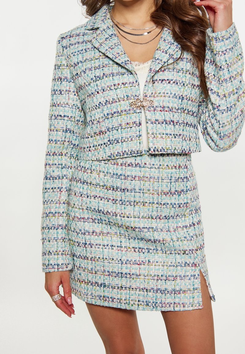 Femme portant une veste courte en tweed multicolore et une mini-jupe assortie avec une fente à l'avant, accessoirisée de colliers en argent superposés et de bagues.