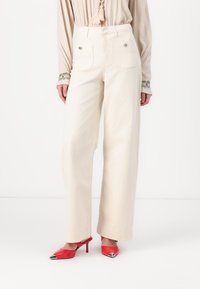 Pantalones de pierna ancha color crema hechos de algodón, con dos bolsillos traseros y detalles en metal plateado, combinados con zapatos rojos de punta. 