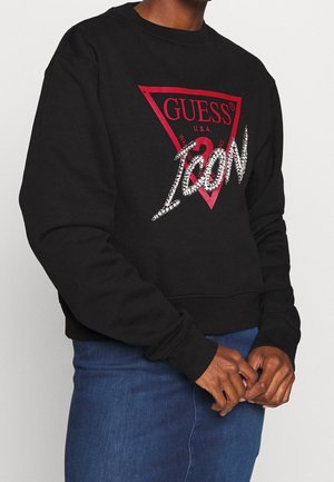 Persoon draagt een zwart sweatshirt met een rood Guess-logo en rhinestone tekst "ICON", gecombineerd met een blauwe spijkerrok.
