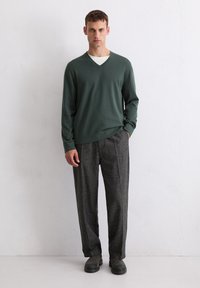 Dunkelgrüner V-Ausschnitt-Pullover aus weichem Stoff, kombiniert mit grauen, strukturierten Hosen aus Tweed und schwarzen Slippern. Steht vor einer hellen Wand.