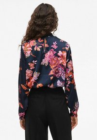 Blusa floreale in blu navy con vivaci fiori rosa, arancioni e viola. Presenta maniche lunghe, polsini arricciati e un dettaglio a chiave sulla schiena.