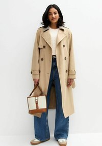 Beżowy płaszcz trench z dużymi klapami, dwurzędowym zapięciem i wyraźnymi guzikami. Noszony na kremowym swetrze z dzianiny i dżinsach o wysokim stanie.