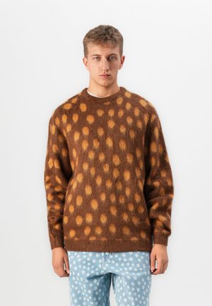 Jonge man in een bruine fuzzy trui met oranje vlekken en lichtblauwe broek met witte stippen, staande tegen een witte achtergrond.