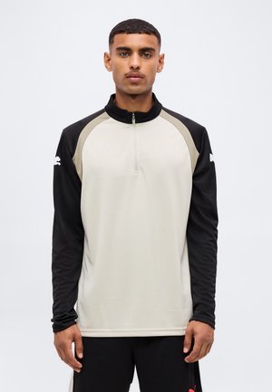 Homme portant un haut de sport beige et noir à manches longues avec une fermeture éclair quart, debout devant un fond clair uni.