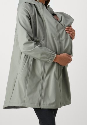 Mujer con una chaqueta gris para porteo de bebés con un bolsillo con cremallera, sosteniendo a un bebé dormido cerca de su pecho.