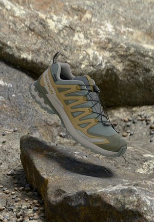 Zapatilla de trail running Salomon en verde claro y beige flotando sobre suelo rocoso con pequeñas piedras dispersas.