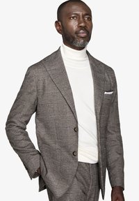 Brun ternet blazer med to knapper, parret med en hvid rullekrave. Har et lommetørklæde og struktureret stofdetaljer.