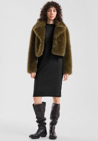 Veste cropped en fausse fourrure olive clair sur une robe pull noire, associée à des bottes en cuir noires à talons hauts. Apparence texturée et chaleureuse.