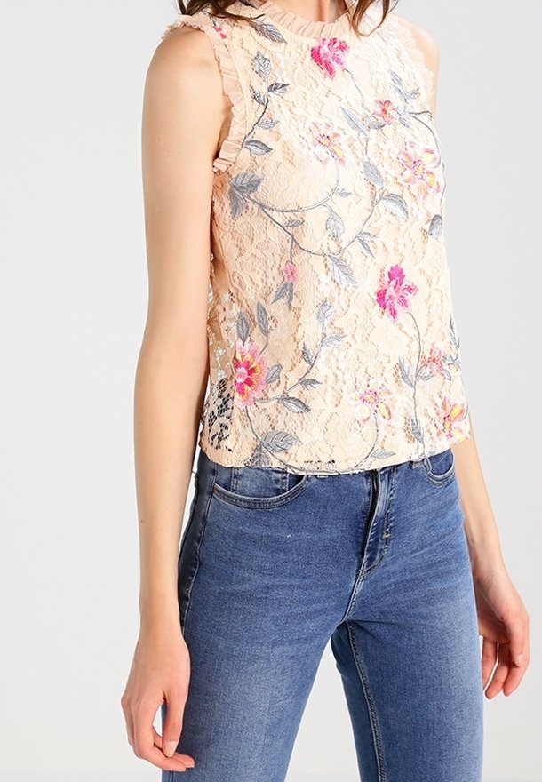 Top en dentelle beige sans manches avec broderies florales roses et violettes porté avec un jean en denim bleu sur une personne debout.