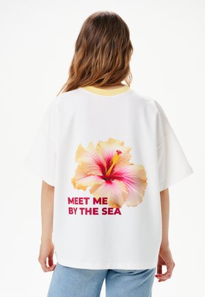 Osoba o falowanych brązowych włosach nosi białą, oversize koszulę z dużym różowo-żółtym kwiatem hibiskusa i tekstem "MEET ME BY THE SEA" na plecach.
