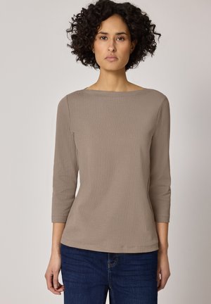 Vrouw met kort, krullend haar, gekleed in een beige geribbelde lange mouwen top en donkere blauwe jeans, staand tegen een neutrale achtergrond.