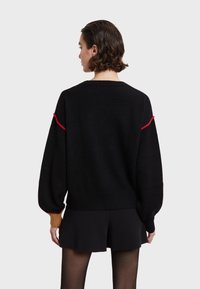 Maglione nero lavorato a maglia con una vestibilità ampia, caratterizzato da accenti rossi sulle spalle e polsini a coste. La texture appare morbida e accogliente.