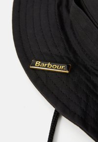 Chapeau seau en tissu noir avec un bord cousu et un petit badge rectangulaire doré et noir de la marque Barbour sur le côté.
