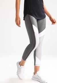 Nainen pukeutuneena harmaisiin ja valkoisiin väri-blokattuihin legginseihin ja valkoisiin urheilukenkiin, seisoo yksinkertaista valkoista taustaa vasten.