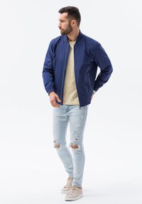 Marinblå bomberjacka, lätt material, dragkedjeslutning, två sidofickor. Bärs med beige t-shirt, ljusblå slitna jeans och beige sneakers.