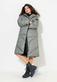 Longo cappotto piumino grigio con cuciture diagonali orizzontali, cappuccio e zip frontale. Abbinato a stivaletti neri con lacci e suola spessa.