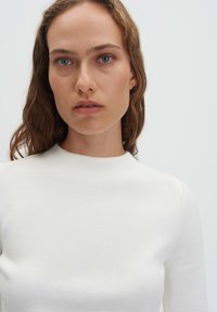 Frau mit welligen braunen Haaren und blauen Augen, die ein schlichtes weißes Langarmshirt vor einem hellgrauen Hintergrund trägt.