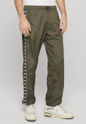 Mann iført olivengrønne treningsbukser med hvite stripes på siden og hvite joggesko, stående mot en enkel lys bakgrunn.