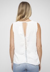 Witte mouwloze top met getextureerde polka dots, voorzien van een centrale opening aan de achterkant met kantaccent. Gladde stof met een losse pasvorm.