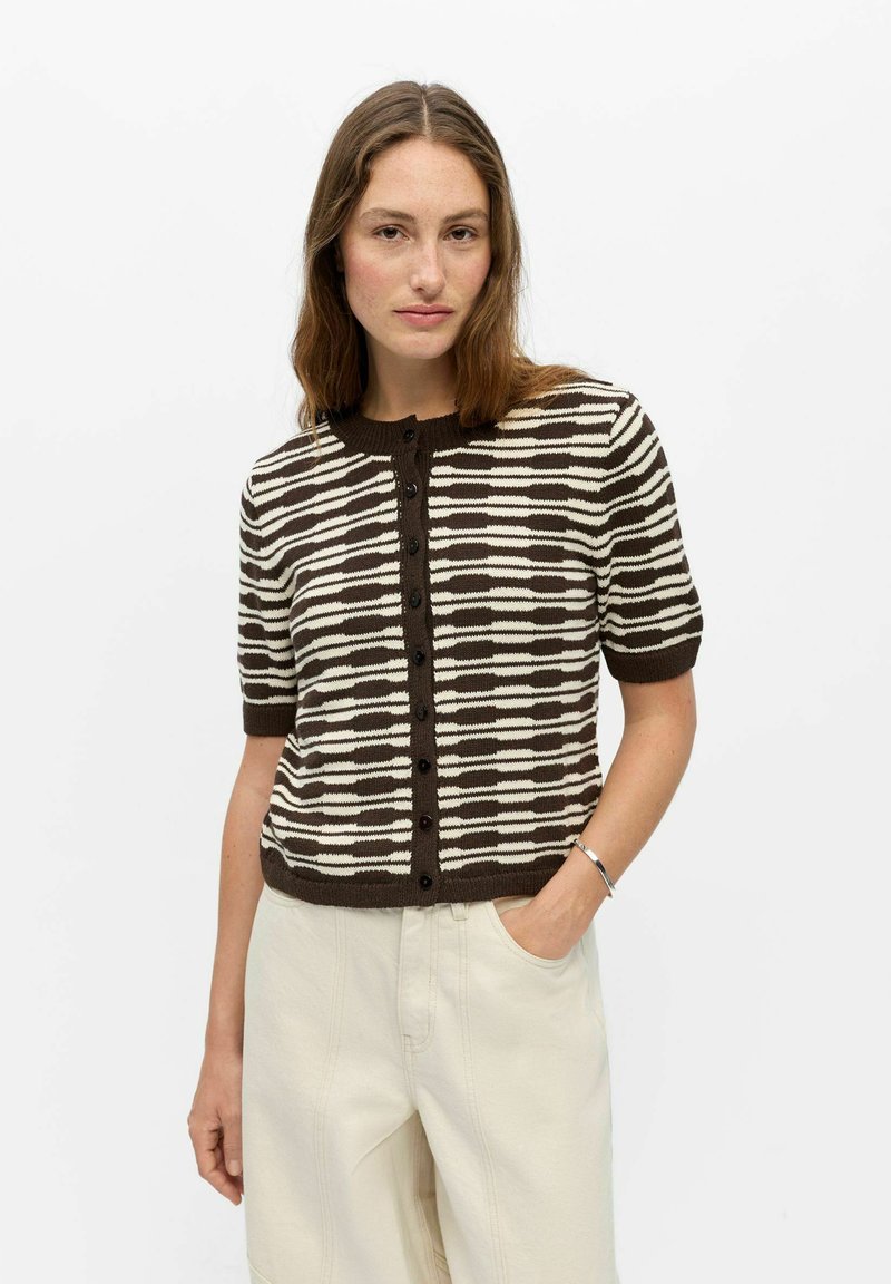 Femme aux longs cheveux bruns portant un cardigan boutonné à manches courtes rayé marron et blanc et un pantalon beige clair avec une main dans la poche.