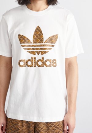 Personne portant un T-shirt blanc avec un grand logo Adidas marron à motifs et un short assorti visible en bas.