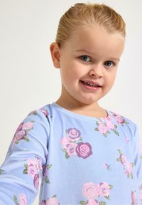 Młoda dziewczyna o blond włosach, ubrana w jasnoniebieską bluzkę z różowo-fioletowymi kwiatowymi wzorami, uśmiechająca się na jednolitym białym tle.