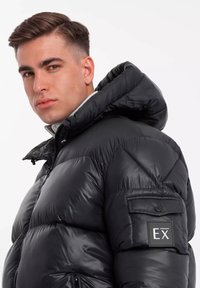 Zwarte pufferjack met een geborduurd ontwerp, een opvallende capuchon, een rits aan de voorkant en een patch op de linker mouw met "EX." Glad, glanzend textuur.