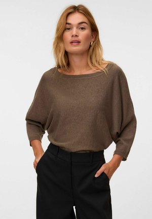 Blond kvinde iført en løs brun sweater og sorte bukser med hænderne i lommerne mod en ensfarvet hvid baggrund.