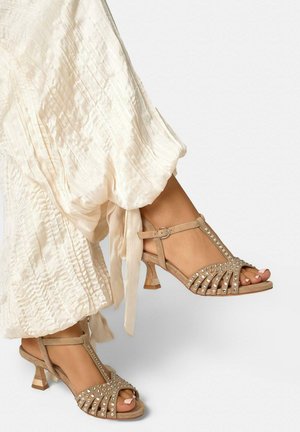 Voeten in beige T-strap sandalen met hak en strassteentjes, gecombineerd met losse, lichtgekleurde, gestructureerde broeken die bij de enkels zijn gestrikt.