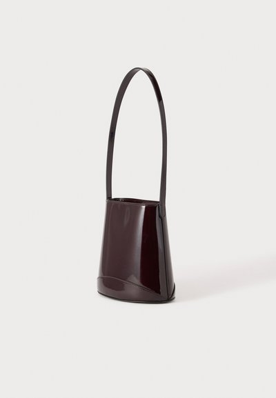 Petit sac seau brun foncé brillant avec une longue bandoulière simple, debout sur un fond clair uni.