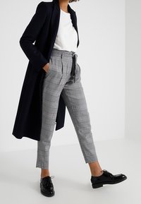 Kvinna i svart långkappa, vit topp, grå rutig hög midja ankelbyxor och svarta snörskor, stående med händerna i fickorna.