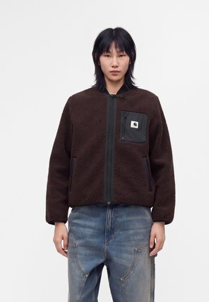 Personne portant une veste polaire marron foncé zippée avec une poche poitrine noire et un jean bleu ample sur un fond blanc uni.