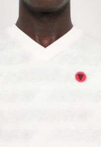 Hvid v-hals sweater med et subtilt struktureret mønster og et rødt broderet badge med et solstråle-design på venstre bryst.