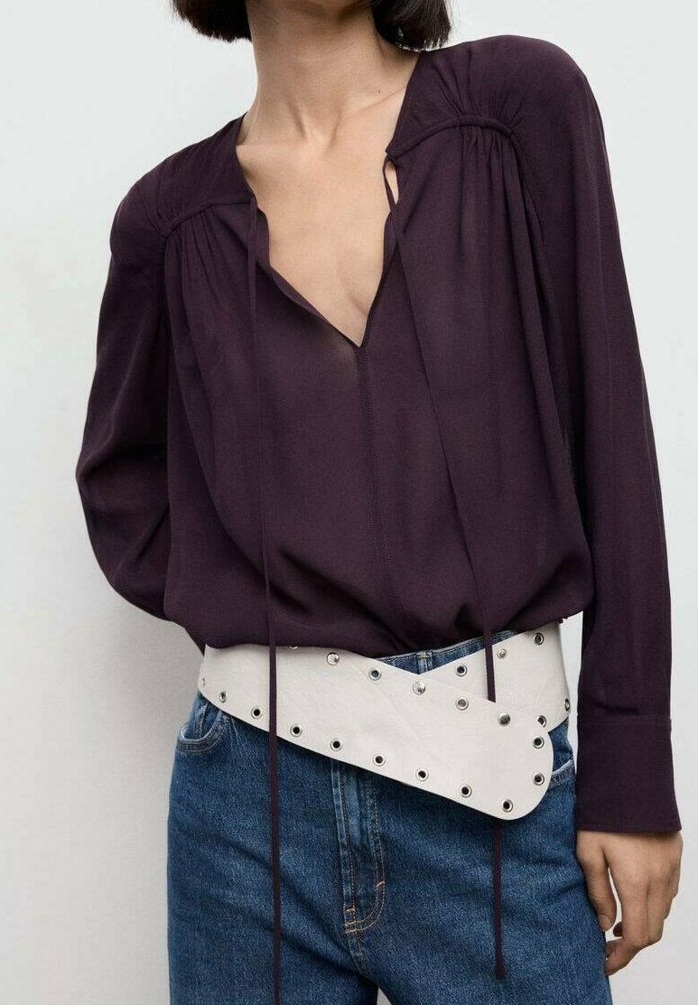 Femme portant un chemisier à manches longues violet foncé rentré dans un jean bleu avec une large ceinture blanche ornée d'œillets métalliques.