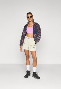Mehrfarbige Fleecejacke mit einer Reißverschlusstasche, leicht lila Cropped-Top, beigefarbene Shorts mit Taschen und schwarze Wanderstiefel.