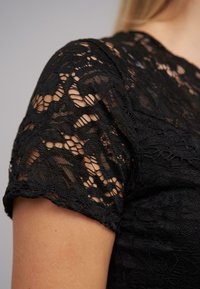 Svart spets topp med korta ärmar, prydd med intrikata blommönster och en semi-genomskinlig textur. Ärmarna når till armbågen och visar detaljerat spetssöm.