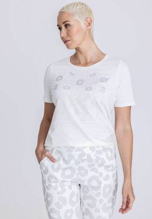 Vrouw met kort blond haar die een wit shirt met korte mouwen draagt met een subtiel patroon en witte broek met lichtgrijze luipaardprint.