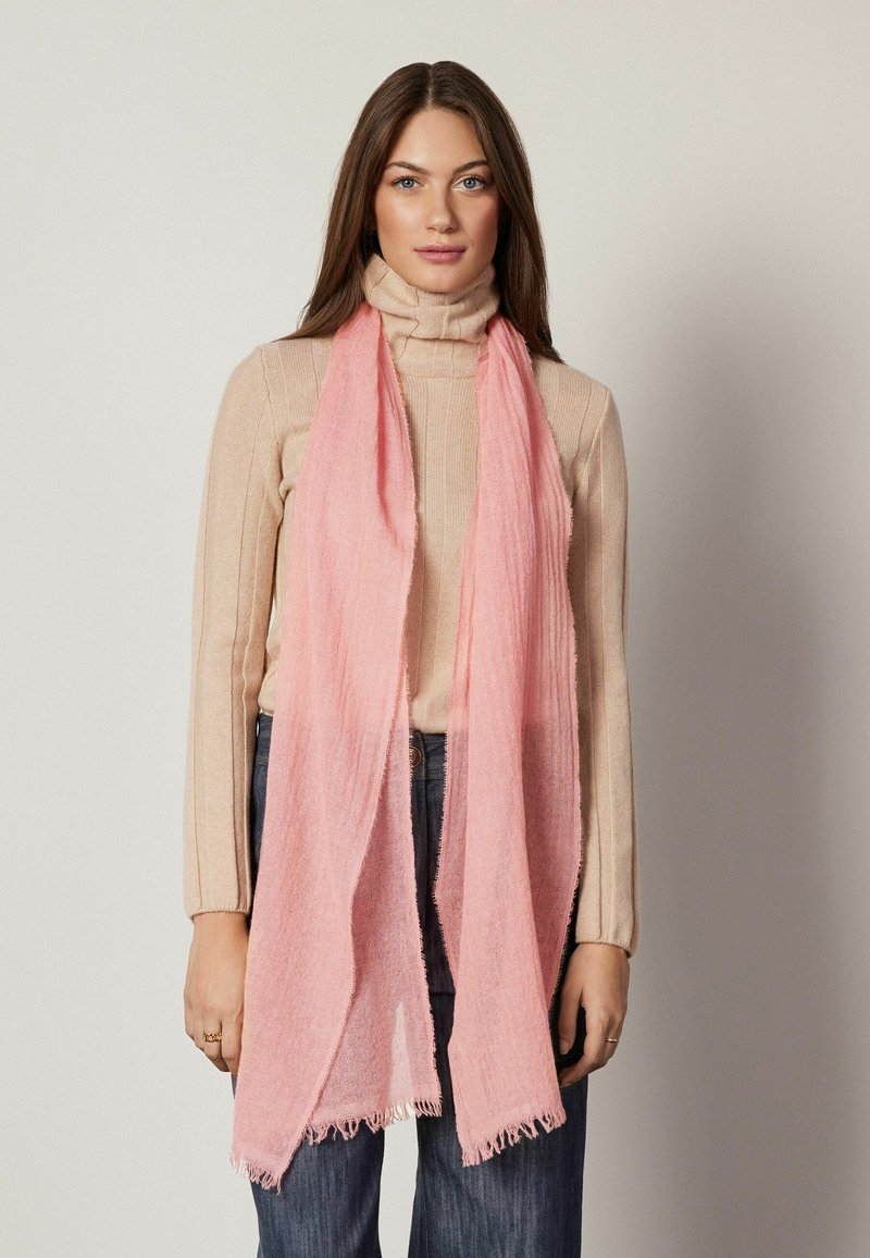 Écharpe rose en tissu léger et texturé avec des bords frangés, superposée à un pull col roulé beige et portée avec un jean foncé.