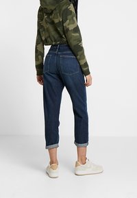 Džínové kalhoty s vysokým pasem, ohrnutými lemy a zadními kapsami. Kombinované se zelenou maskáčovou crop mikinou a bílými teniskami.