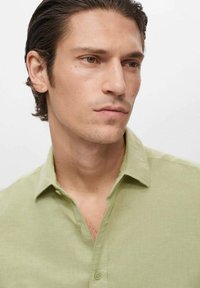 Camisa de lino verde claro con cuello abotonado y mangas cortas, que presenta una textura suave y un brillo sutil.