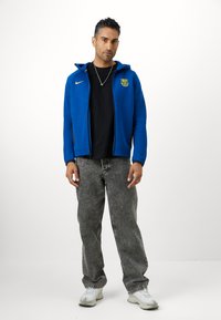 Μπλε φούτερ με φερμουάρ και κουκούλα, συνδυασμένο με μαύρο t-shirt και ξεθωριασμένα γκρίζα τζιν. Λευκά αθλητικά παπούτσια, ελάχιστα κοσμήματα, ουδέτερο φόντο.