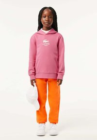Felpa in cotone rosa con logo bianco, abbinata a pantaloni della tuta arancioni brillanti. Sneakers bianche e cappellino bianco tenuto nella mano sinistra.