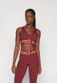 Burgundska športna modra z izrezom, ki ima oranžni pas "PRO NIKE", v kombinaciji z usklajenimi leginami in majhnim belim logotipom.